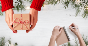 Hands holding a gift box and writing in a notebook surrounded by holiday decor