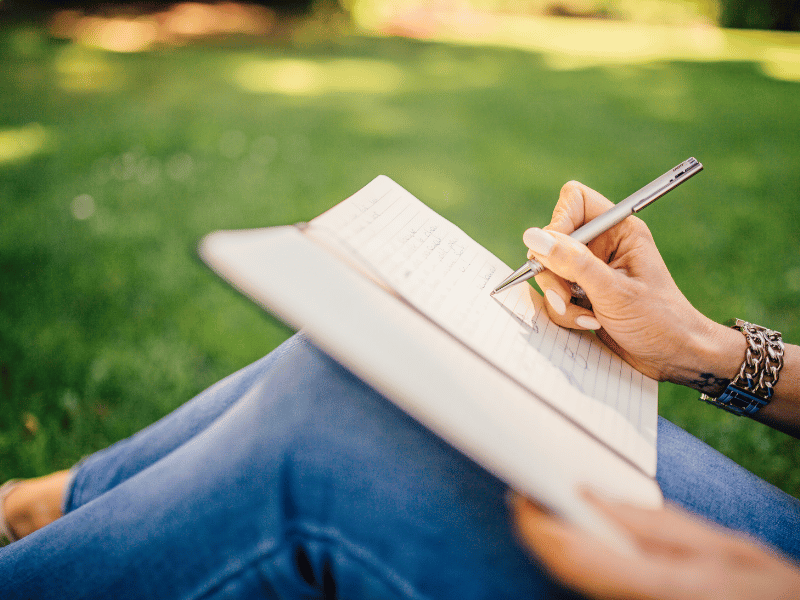 A person sitting on grass, journaling in a notebook with a pen, surrounded by nature.