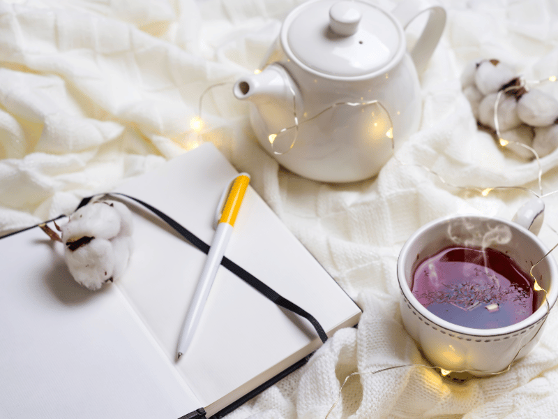 A journal with a pen, teapot, and cup of tea on a soft blanket, symbolizing writing as a mindful and comforting practice.