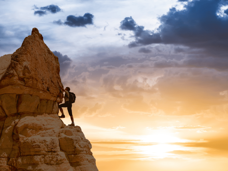 A silhouette of a person climbing a rocky cliff with a radiant sunrise in the background, symbolizing ambition and personal growth.