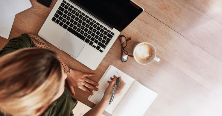 Overhead view of a person writing in a notebook with a laptop and coffee nearby, symbolizing creativity, focus, and accessibility of writing.