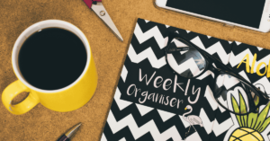 Flat lay of a yellow coffee mug, weekly organizer notebook with chevron pattern, a pair of glasses, and a tablet on a wooden desk