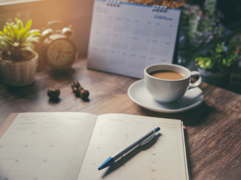 Notebook with a pen, a coffee cup, and a calendar on a wooden desk surrounded by greenery, symbolizing a well-organized workspace