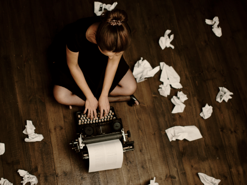 Frustrated writer sitting on the floor surrounded by crumpled paper and a typewriter, symbolizing writer's block.