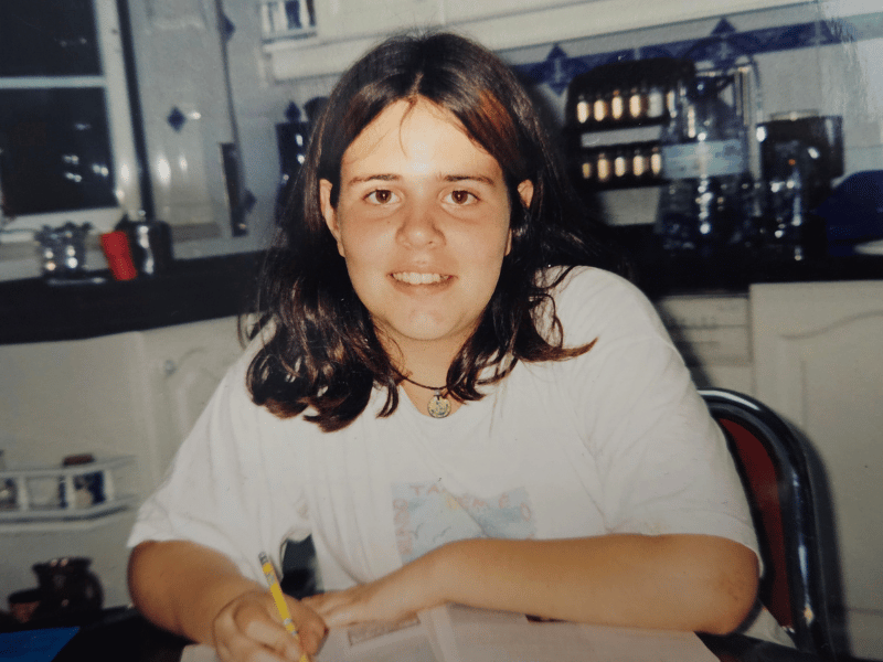 A young girl writing in a notebook at a kitchen table, smiling at the camera.