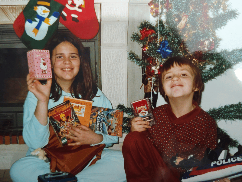 A young girl holding a stack of books on Christmas morning, smiling beside her brother.