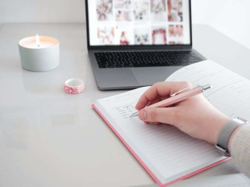 A clean, minimal desk setup with a laptop, a candle, and an open notebook with a pink pen.