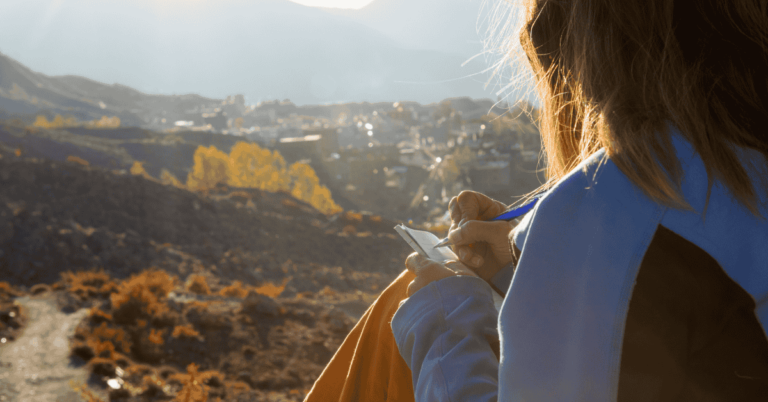 woman writing in a notebook with a scenic view of mountains and a valley in the background, bathed in golden sunlight.