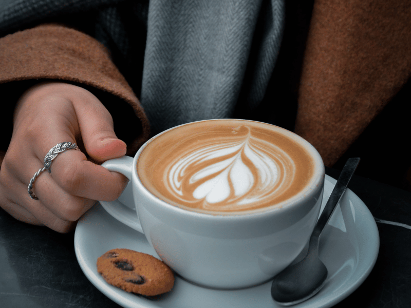 Hand holding a cup of latte with art on the foam, served with a small cookie.