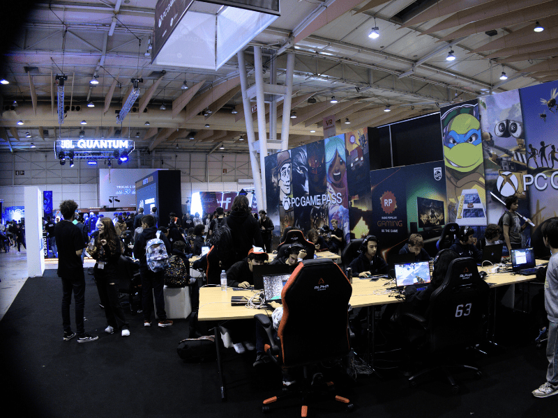 Crowd attending a gaming convention surrounded by brand booths and computers under bright industrial lights.