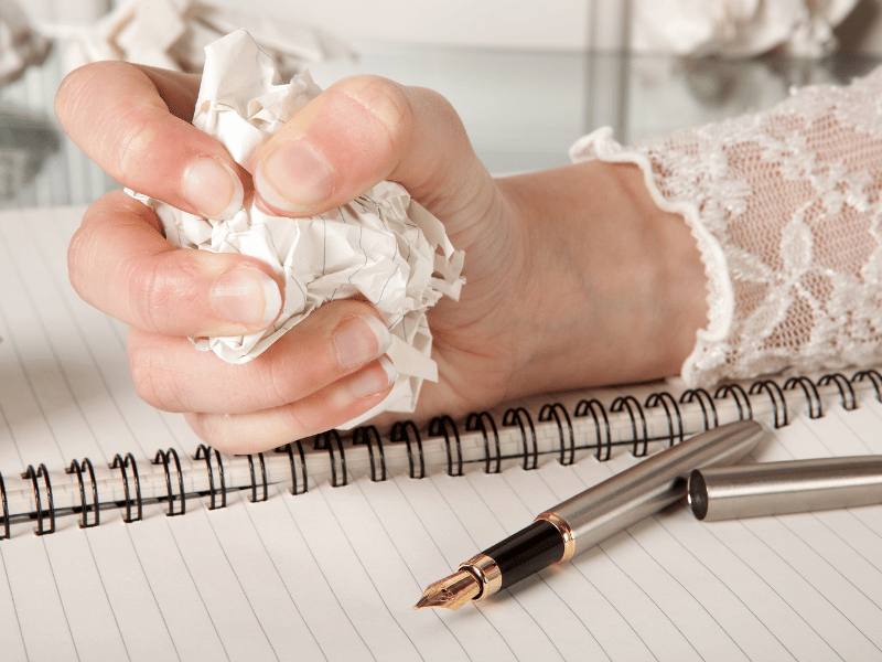 A hand crushes a piece of paper over a notebook and fountain pen, representing frustration and perfectionism in writing.