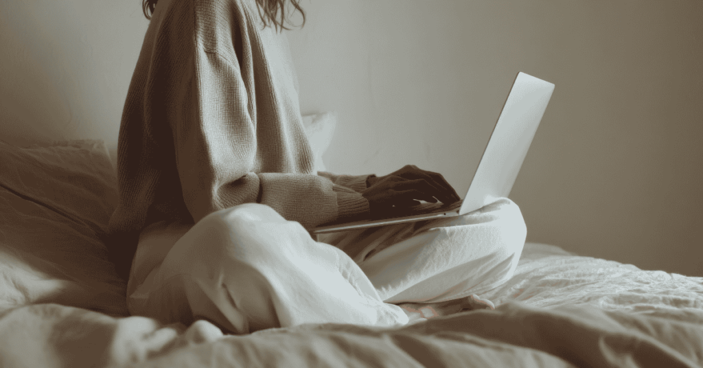 Person sitting on a bed writing on a laptop, representing gentle writing without pressure or rigid workspace.