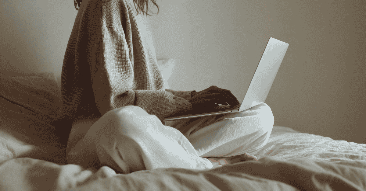 Person sitting on a bed writing on a laptop, representing gentle writing without pressure or rigid workspace.