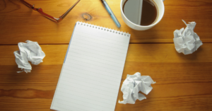 crumpled papers, blank notebbook, scattered pencils and a coffee mug on a wooden desktop.