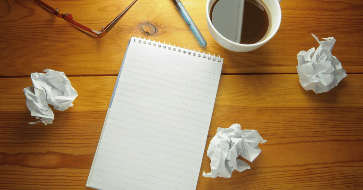 crumpled papers, blank notebbook, scattered pencils and a coffee mug on a wooden desktop.