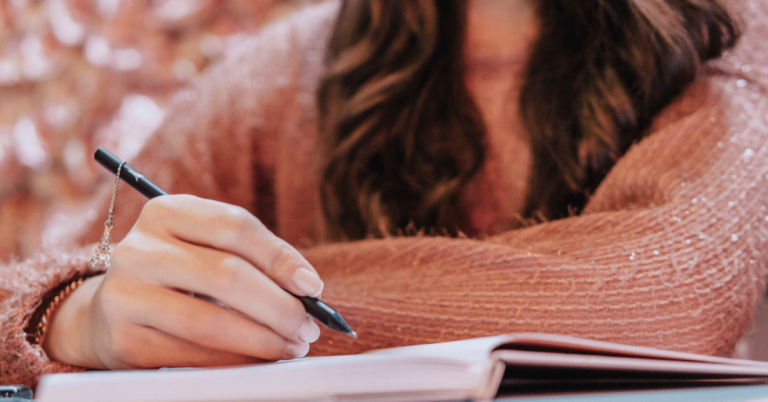 Woman in a pink sweater writing by hand in an open notebook, close-up of her hand holding a pen.